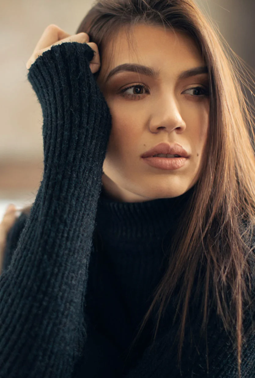 A portrait of a woman with long, brown hair wearing a dark, ribbed turtleneck sweater. She is looking to her right, and her right hand is raised to her head, with her fingers resting near her temple. - Liquid Facelift in Draper, UT