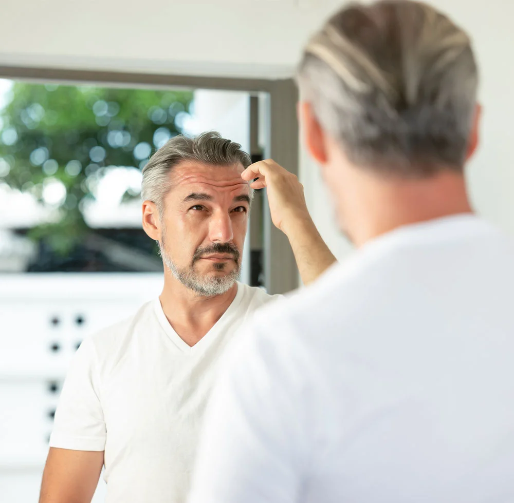 A handsome, middle-aged man with graying hair and a beard, wearing a white t-shirt. He is looking at his reflection in a mirror and raising his right hand to touch his forehead. - Male Brow Reset in Draper, UT
