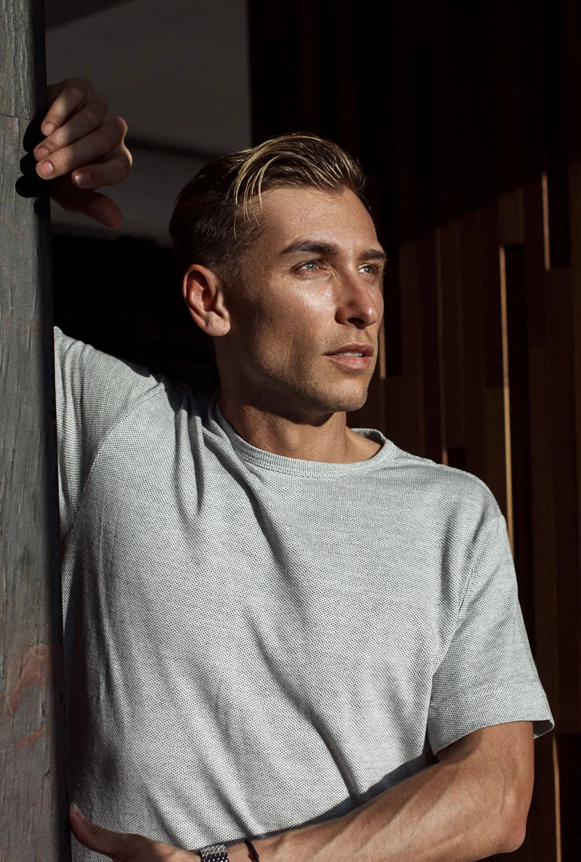 A portrait of a young man with blonde hair, wearing a gray, textured t-shirt. He is leaning against a dark wooden pillar, with his left arm resting on it and his right arm on his hip, looking away to his right. Sunlight is illuminating his face and hair from the right. - Male Brow Reset in Draper, UT