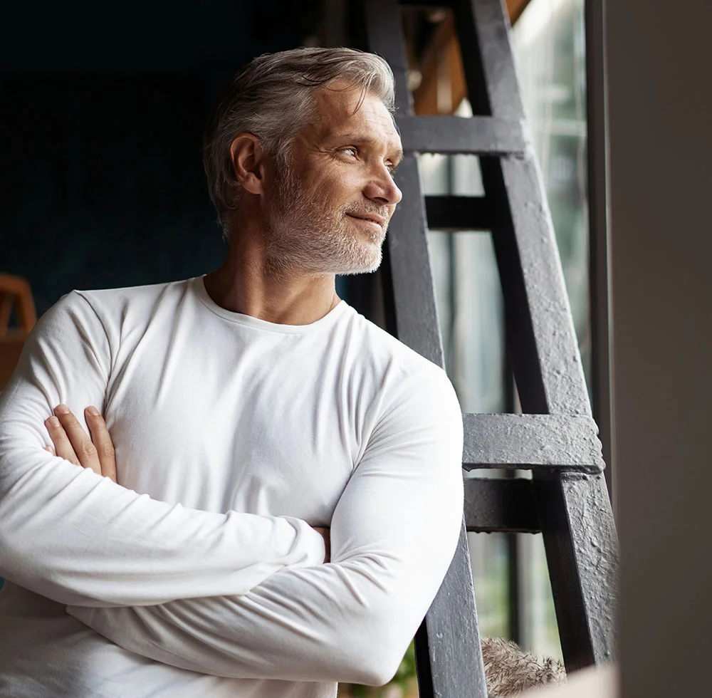 A middle-aged man with gray hair and a beard, wearing a white long-sleeved shirt, standing with his arms crossed. He is looking out a large window to his right with a happy expression on his face. - Male Eyelid Surgery in Draper, UT