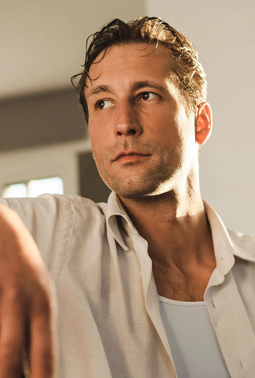 A man with dark, slicked-back hair, wearing a white collared shirt and a tank top underneath, is looking thoughtfully to his left. He is seated with his head slightly tilted, and the light from the sun is on the left side of his face. - Male Eyelid Surgery in Draper, UT