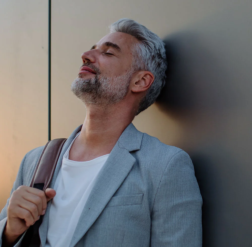A relaxed man with gray hair and a beard, wearing a white t-shirt and a gray sport coat, is leaning against a gray wall with his eyes closed and his head tilted back and to the left. He is holding the strap of a brown bag over his right shoulder. - Male Facelift and Neck Lifts in Draper, UT