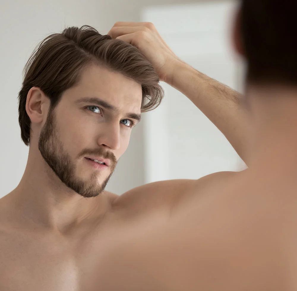 A man with a neatly trimmed beard and brown hair is looking directly at the camera, with one hand running through his hair. His blue eyes are prominent, and his bare shoulder and chest are visible, suggesting a close-up, personal shot. The background is a soft, light color. - Male Hair Restoration in Draper, UT