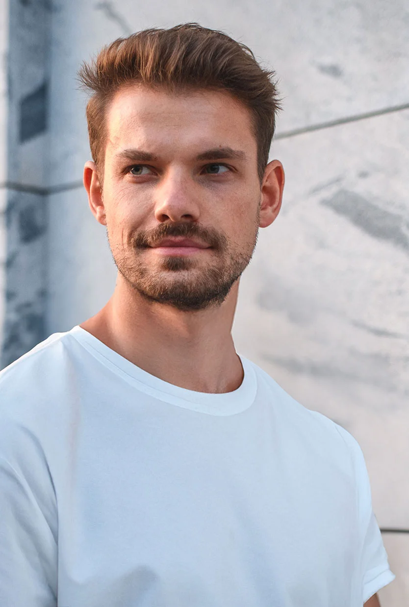 A well-groomed man with a short beard and stylishly coiffed hair looks thoughtfully to the right, standing against a modern building with gray and white marble-like panels. He is wearing a plain white crew-neck t-shirt. - Male Hair Restoration in Draper, UT