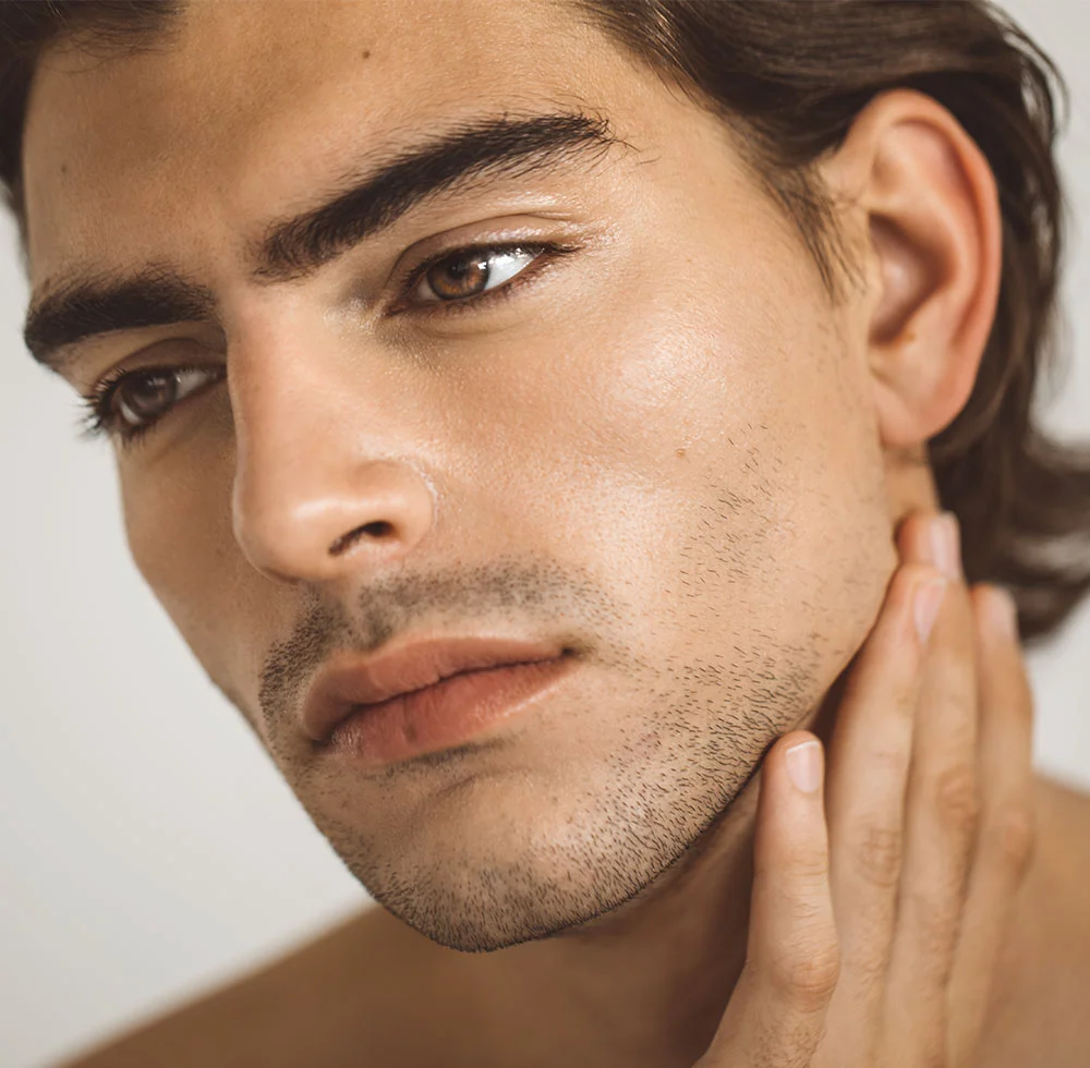 Close-up of a light-tan man with short brown hair and a sparse dark beard. His light brown eyes look slightly downward to the left. A hand rests on his neck and jaw, emphasizing his smooth, even skin under soft gray lighting. - Male Injectables in Draper, UT