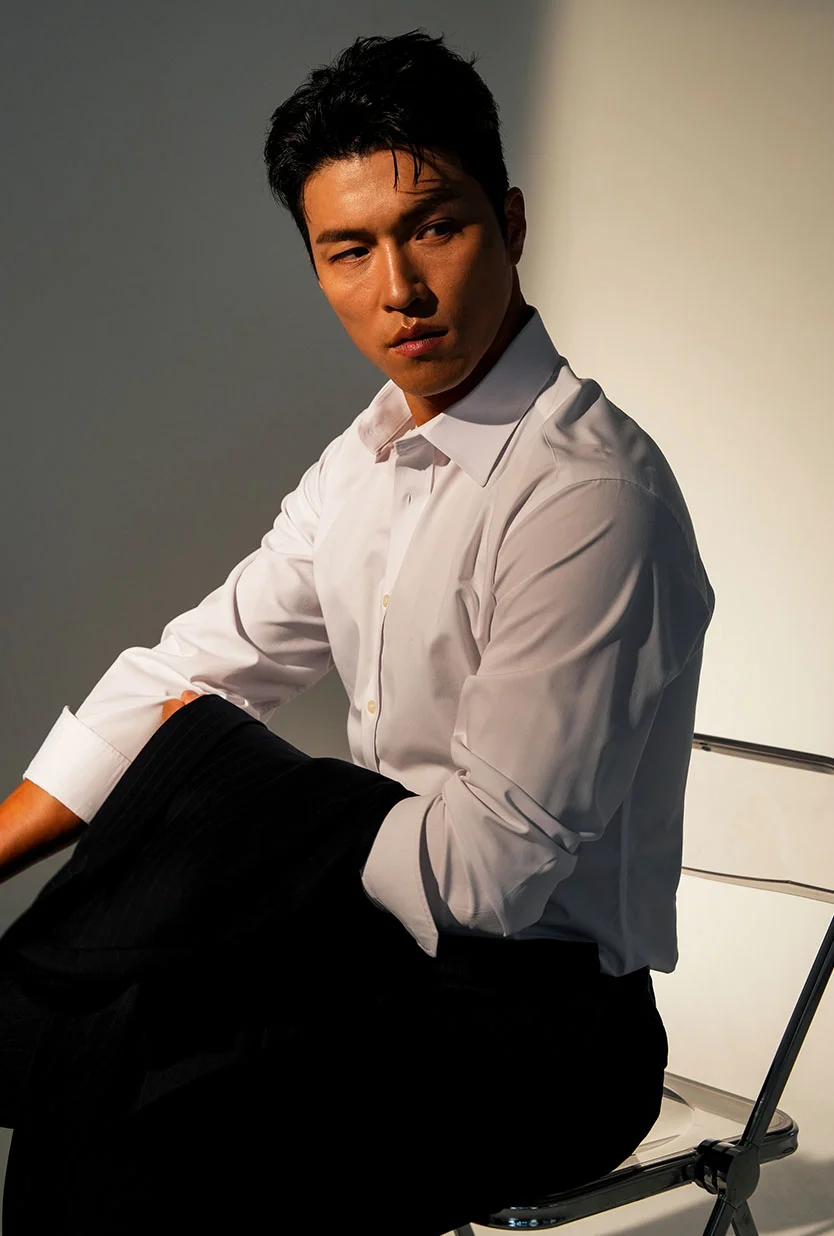 A light-skinned man with short dark hair sits angled toward the camera in a chair. He wears a light off-white button-up shirt with rolled sleeves and dark straight-cut pants. The muted gray-white studio background and soft lighting create an elegant, understated mood. - Male Injectables in Draper, UT