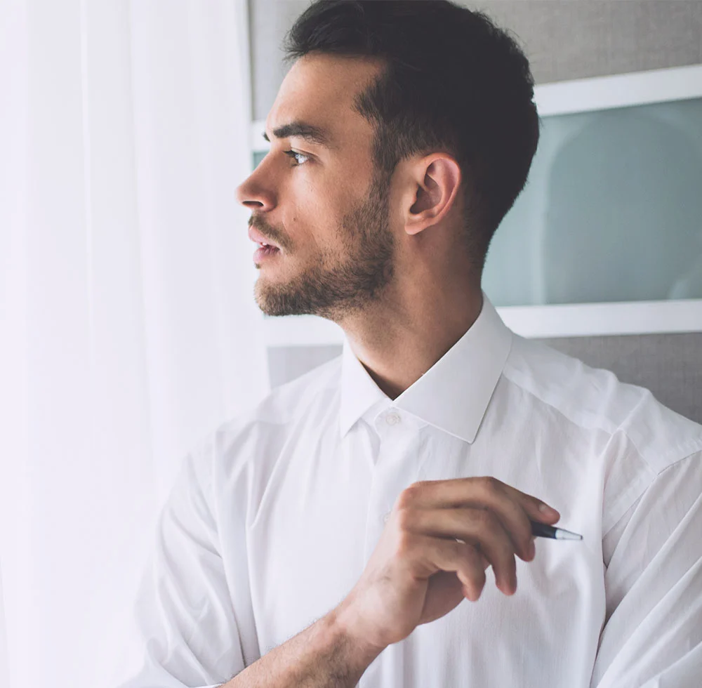 Close-up profile of a light-skinned man with short dark hair and a light beard. He wears a white button-down shirt and holds a black pen near his chest, looking thoughtfully to his right. The background shows a blurred light-colored wall with sheer curtains on the left and a gray-toned wall with a framed piece on the right. - Male Jawline Contouring in Draper, UT