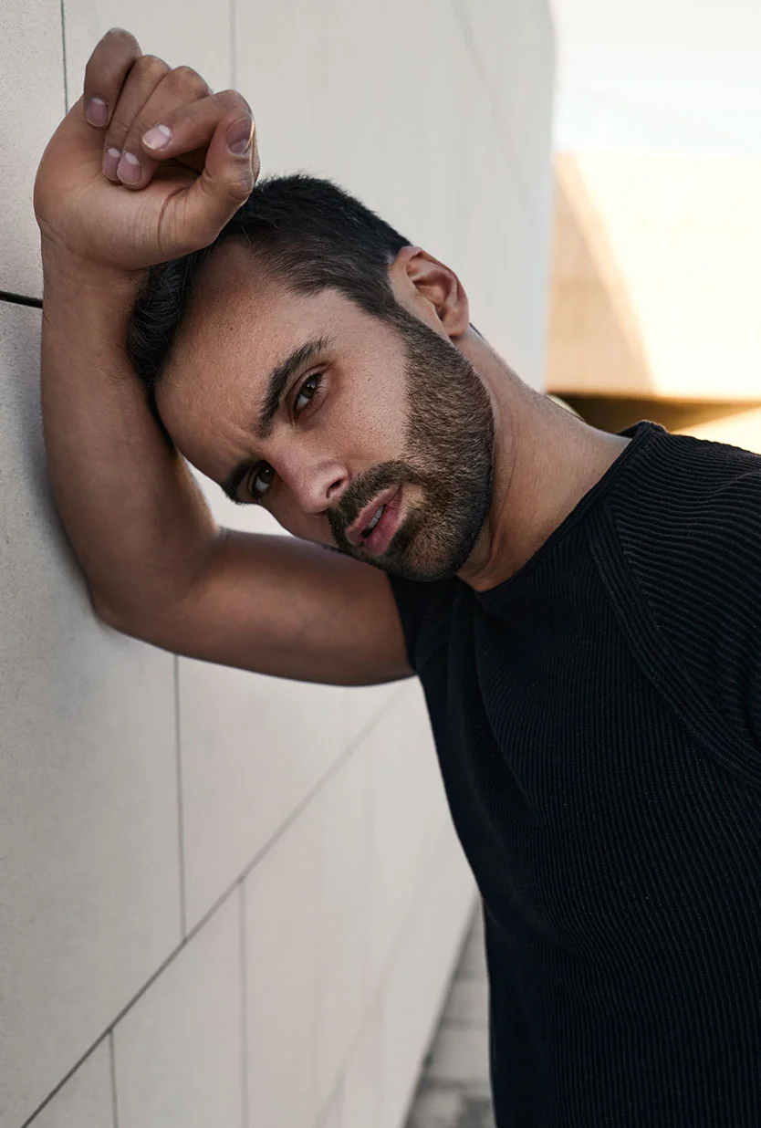 A man leans against a light gray tiled wall, resting his head on his right arm. He has dark brown hair, a short dark beard, and wears a fitted black ribbed t-shirt. He looks at the camera with a neutral expression. Natural light casts subtle shadows, and a light-colored building is faintly visible in the background. - Male Jawline Contouring in Draper, UT