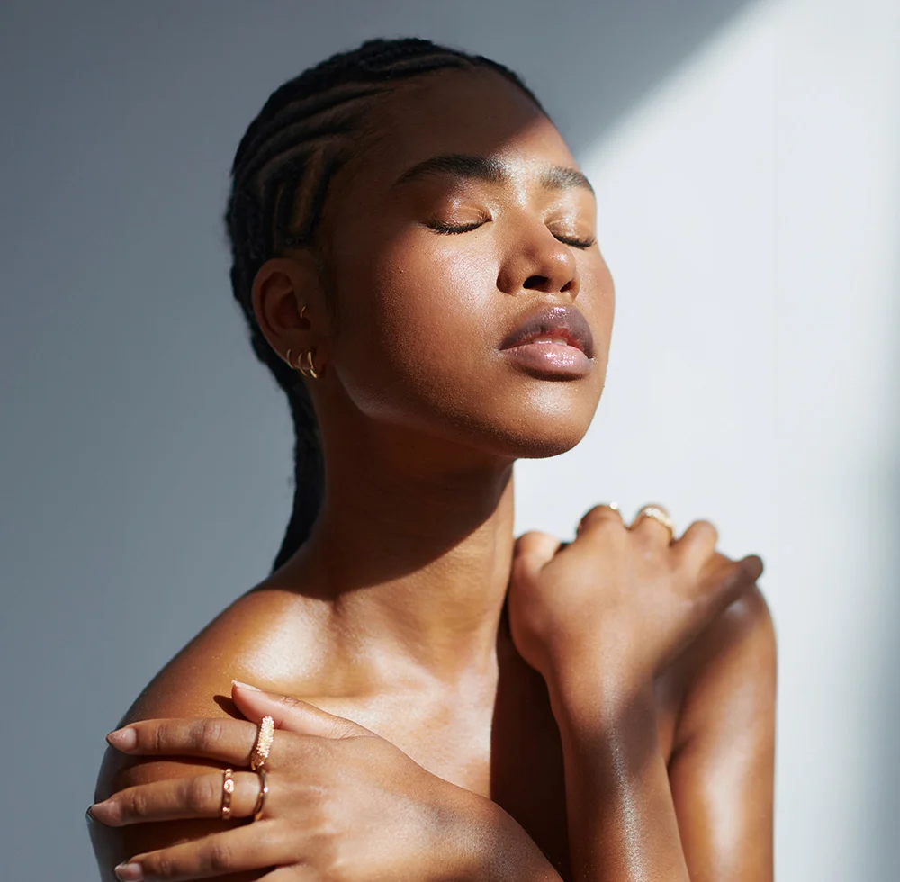 Close-up portrait of a dark-skinned woman with intricate braids. Her eyes are closed, lips light pink, and sunlight softly highlights her smooth, radiant skin. She wears small gold-toned rings on her fingers, resting on her forearms. The background is light gray, and the lighting emphasizes her natural beauty. - Microneedling and RF Microneedling in Draper, UT