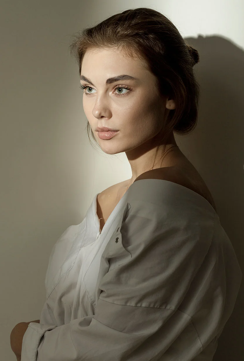 Portrait of a young woman with light brown hair in a bun, shown in three-quarter profile. She gazes thoughtfully to her right, with light hazel eyes and smooth, light-toned skin. She wears a loose, grayish-white long-sleeved shirt, and the off-white background with soft shadows gently highlights her face and upper body. - Microneedling and RF Microneedling in Draper, UT