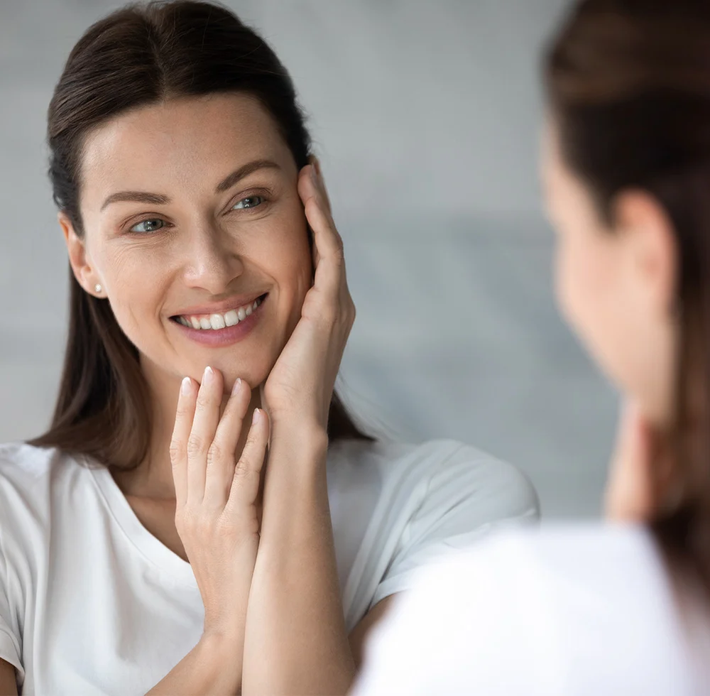A young woman with long, dark hair looks intently at her reflection in a mirror, her face framed by her hands as she gently touches the skin around her eyes, possibly examining or applying a product. She is wearing a simple white tank top, and the background is a soft, blurred reflection, creating a sense of intimacy and focus on her skincare routine. - Mid Facelift in Draper, UT