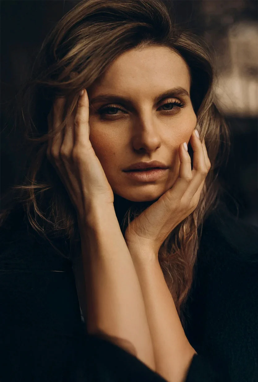 Close-up portrait of a woman with shoulder-length wavy brown hair. She has light-brown skin and a contemplative expression, looking directly at the viewer. Her hands gently frame her face at the temples. She wears a dark, out-of-focus top, and the softly blurred dark background highlights her face with soft, diffused lighting. - Mid Facelift in Draper, UT