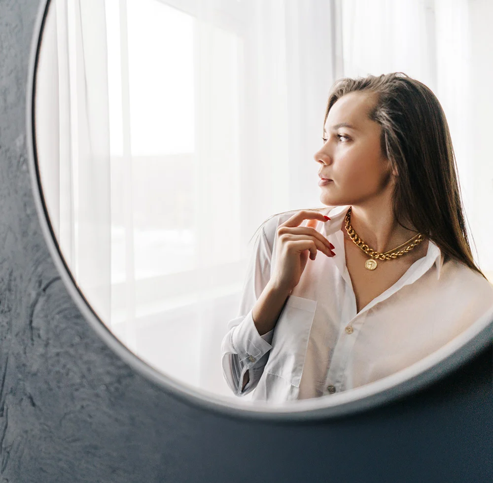 A young woman with long brown hair looks out a window, seen from the side. She wears a white button-down shirt and a gold medallion necklace, with one hand touching her collar. Soft light filters through sheer curtains, and a round grayish-taupe mirror reflects her and the surroundings, creating a calm, reflective mood. - Neck and Chin Liposuction in Draper, UT