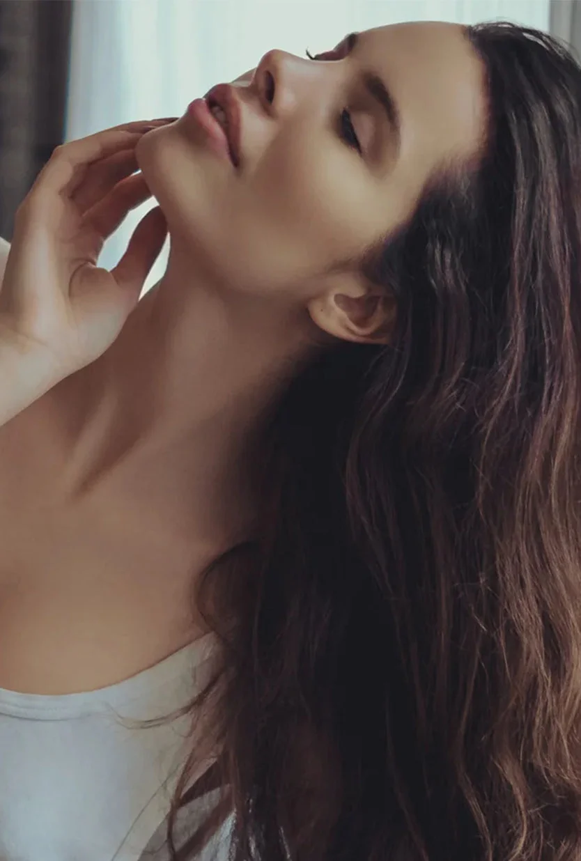 A woman with long, dark brown wavy hair is shown from the mid-chest up in a soft, side-facing pose. Her eyes are closed, head tilted back, and one hand gently touches her neck. She wears a light-colored tank top. The background features a light gray-beige interior with a window and sheer curtains, softly lit to create a calm, serene mood. - Neck and Chin Liposuction in Draper, UT