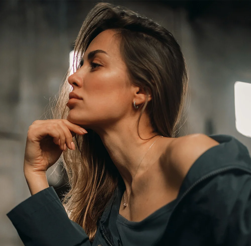 A woman with long, light brown hair is shown in profile from the mid-chest up. Her head is slightly tilted, hand resting on her chin, with a thoughtful expression. She wears hoop earrings, a thin necklace, and a dark-gray or navy-blue top. The dark, textured background is softly illuminated, casting subtle shadows that highlight her facial features. - Neck Lift Recovery in Draper, UT