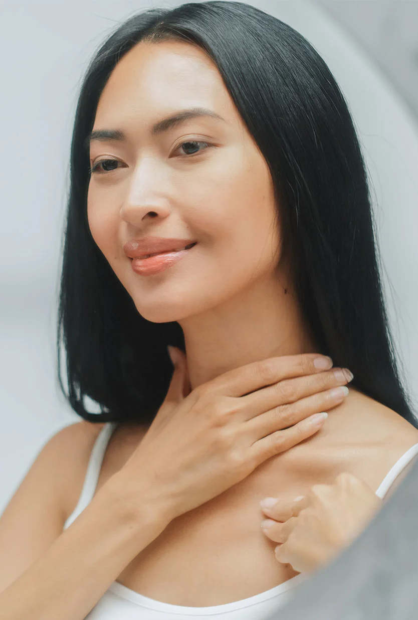 A woman with long, dark hair is looking at her reflection. She has light skin and a soft smile. Her hands are gently placed on her neck and upper chest, likely applying or checking something. She is wearing a white tank top. The background is a light gray, out of focus. The image appears to be of a woman checking her skin. - Neck Lift in Draper, UT
