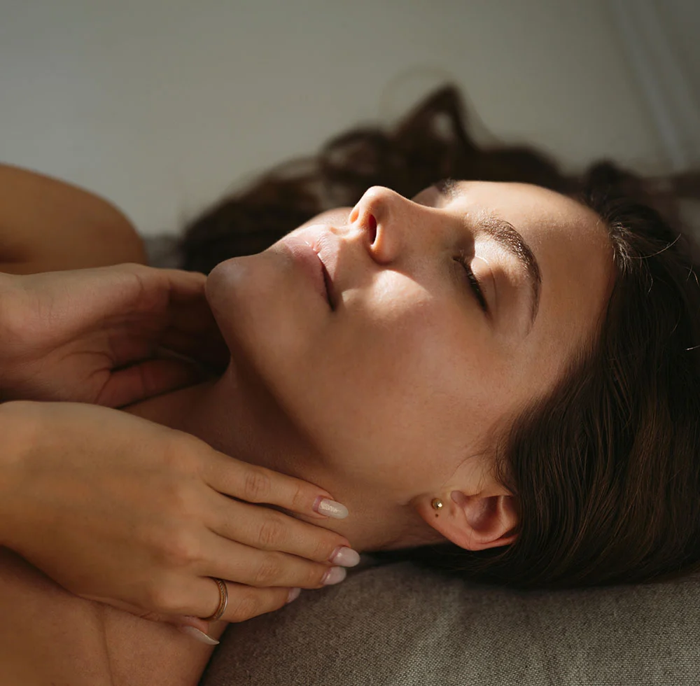 A close-up of a woman lying on her side on a light brown-gray surface. Her dark brown hair cascades over her shoulders, and her eyes are closed in a serene, resting expression. Her hands gently touch her neck and collarbone. She wears small gold earrings and a faint ring, with subtle light-colored nail polish. Soft natural light highlights her face and upper body, while the background remains softly out of focus. - Pico Laser in Draper, UT