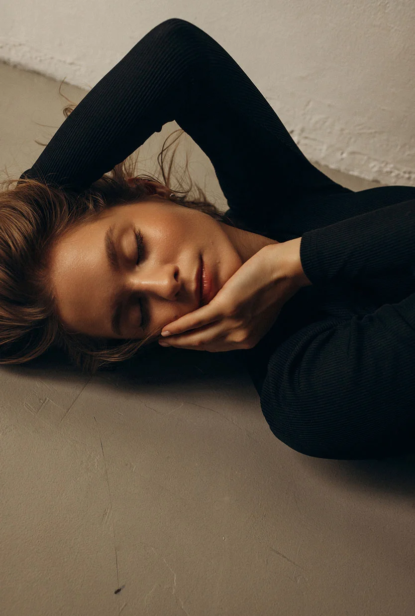 A young woman with light brown hair and a light tan complexion lies on a taupe-colored floor. Her eyes are closed, and her head rests gently on her hands. She wears a long-sleeved, ribbed black top, with the sleeves draped loosely. Soft, diffused light highlights her face and shoulders, while the subtly textured floor adds depth. The overall mood is serene and contemplative. - Picro Laser in Draper, UT