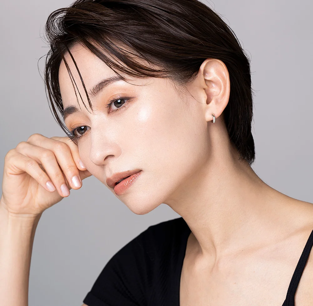 A close-up portrait of a light-skinned woman with short dark brown hair and a luminous complexion. She wears subtle makeup with peachy blush and a rosy lip, along with small silver hoop earrings. Her right hand rests gently on her face as she gazes slightly downward. She is dressed in a black fitted top against a plain light gray background. - PRP PRF in Draper, UT