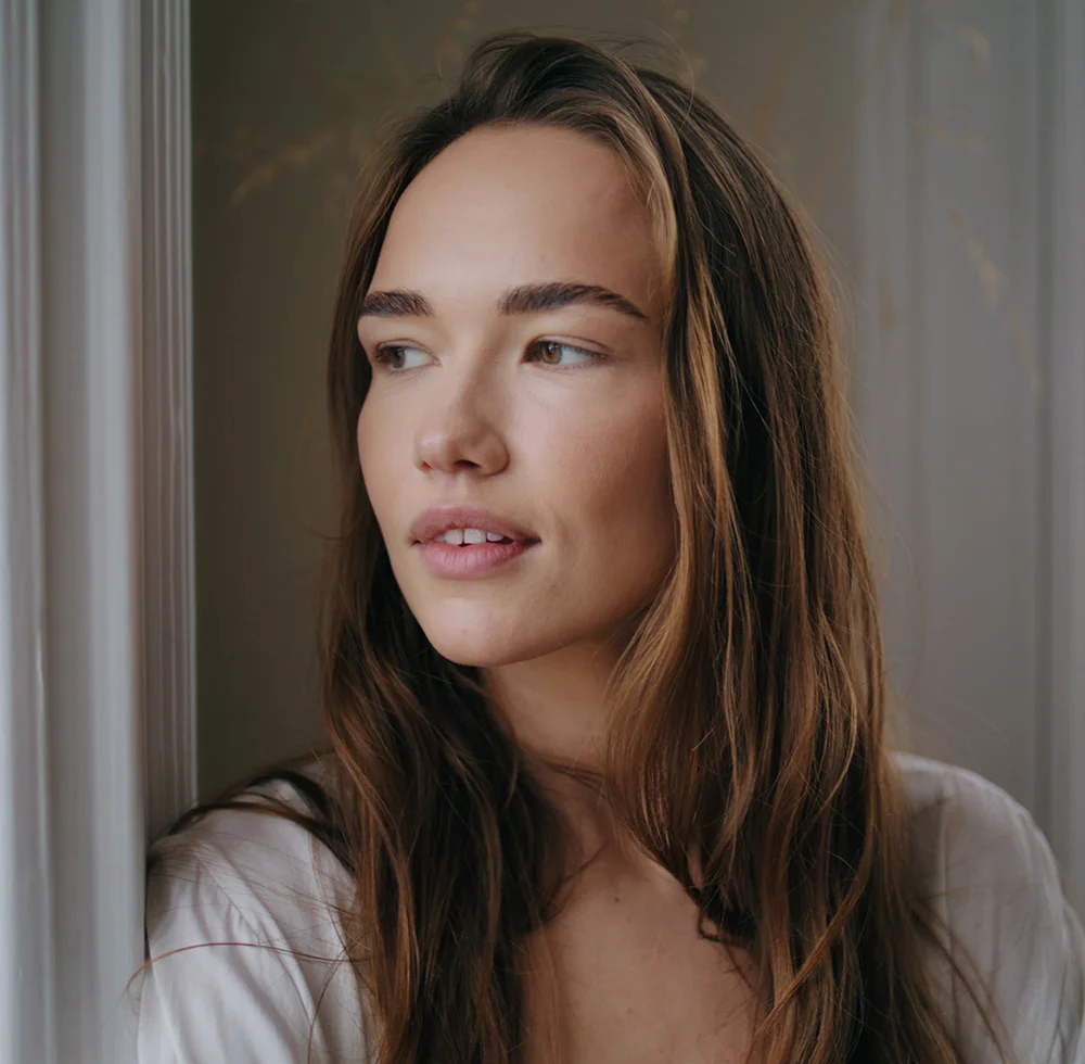 A woman with shoulder-length light brown hair leans against a window frame, gazing to her right with a contemplative expression. She wears a light beige top, and soft indoor lighting highlights her smooth, light-toned skin. The background is a neutral taupe-gray wall with a light window frame, slightly blurred at the edges to keep focus on her face and upper body. - Ptsosis Droopy External Eyelid Surgery in Draper, UT