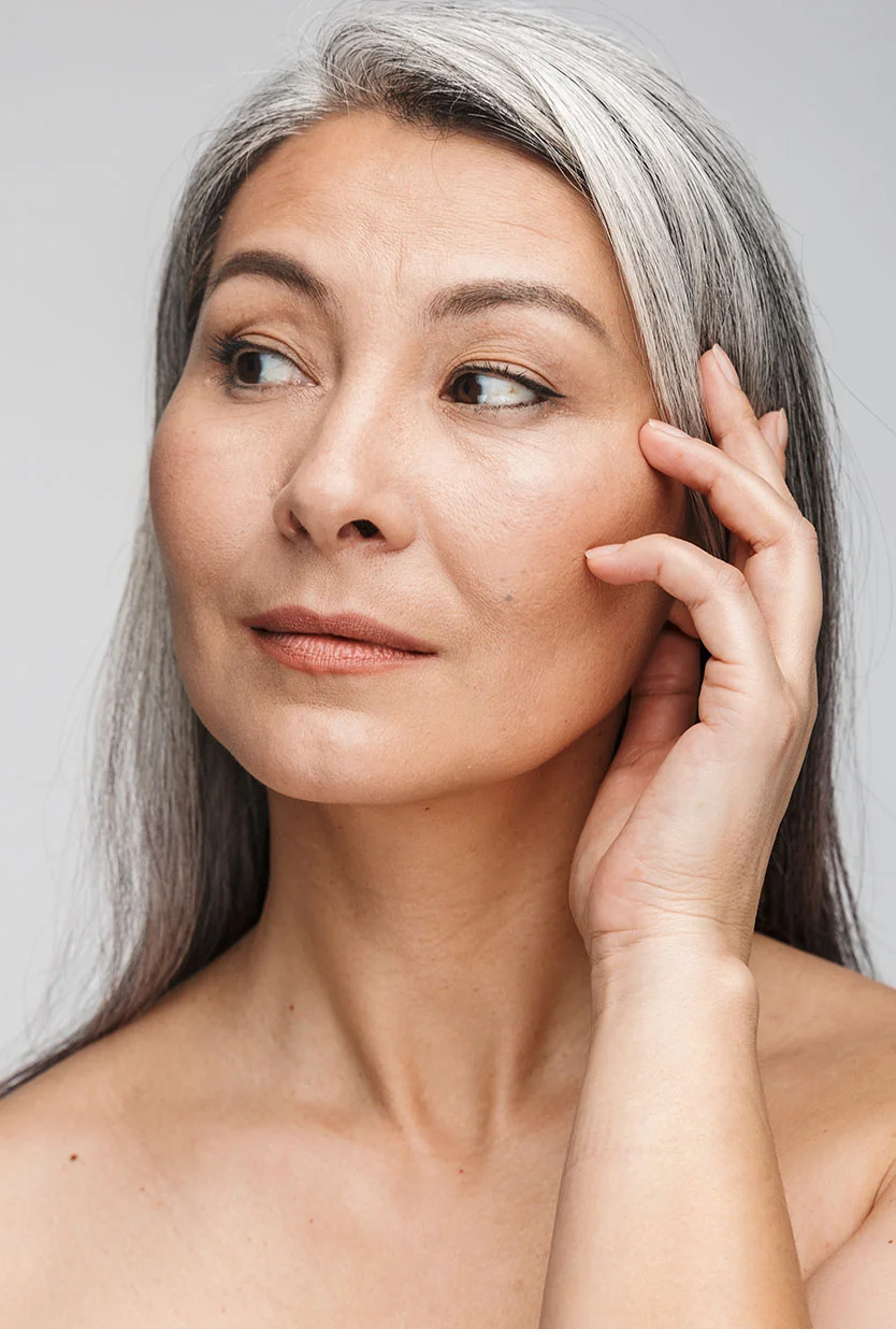 A beautiful woman with long, salt-and-pepper hair, featuring prominent gray strands, looks down softly with a serene expression. She is touching her hair near her ear with one hand. Her shoulders are bare, suggesting a natural or minimalist setting, against a light, plain background. - Ptsosis Droopy External Eyelid Surgery in Draper, UT