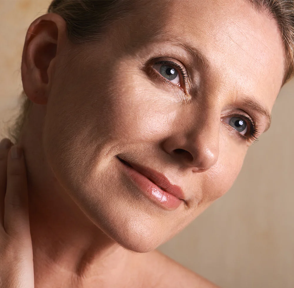 A close-up portrait of a middle-aged or older light-skinned woman shows her face and upper neck. She wears pinkish-nude lipstick and has fine lines on her forehead, eyes, and cheeks. Her light-colored eyes, smooth skin with a subtle glow, and slight serene smile are highlighted against a soft beige background with gentle lighting and shadows. - Quad Blepharoplasty in Draper, UT