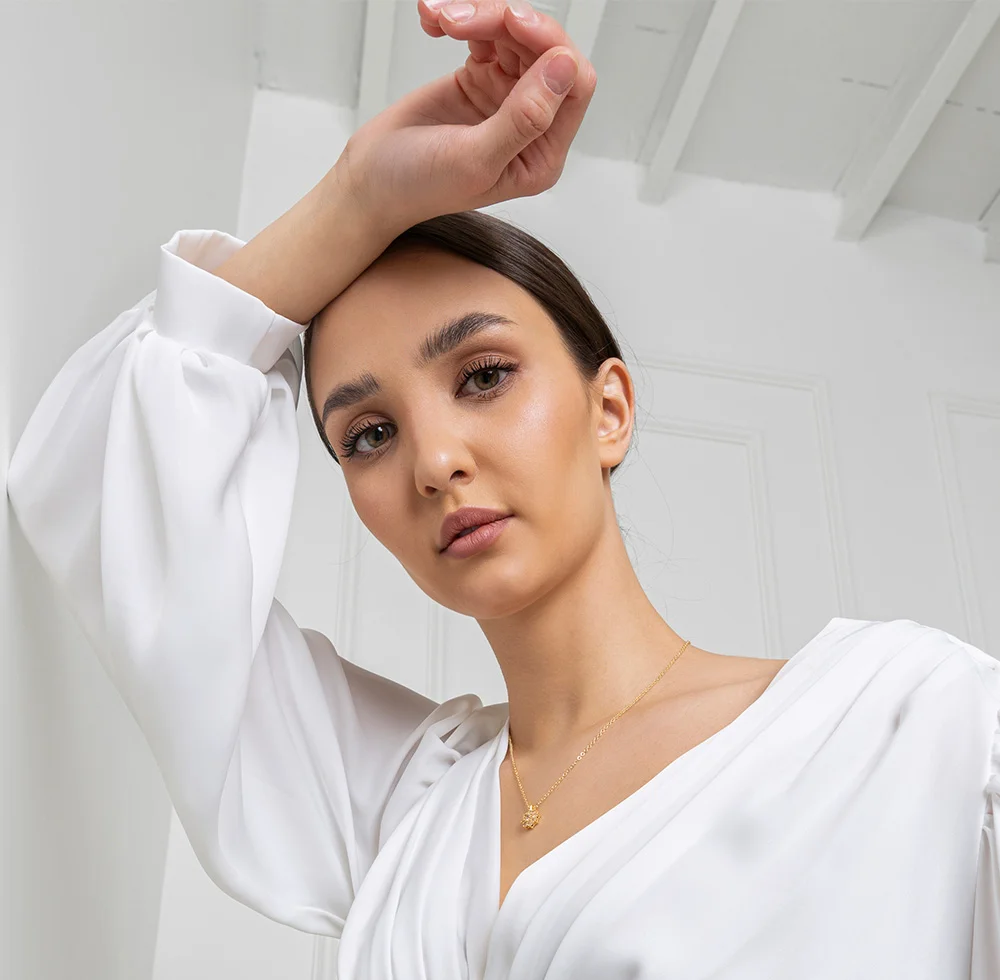 A woman gazes calmly at the viewer, wearing a flowing white blouse with puff sleeves and a gold pendant necklace. Her dark brown hair is pulled back, set against a softly lit cream background that highlights the garment’s elegance. - Revision Eyelid Surgery in Draper, UT