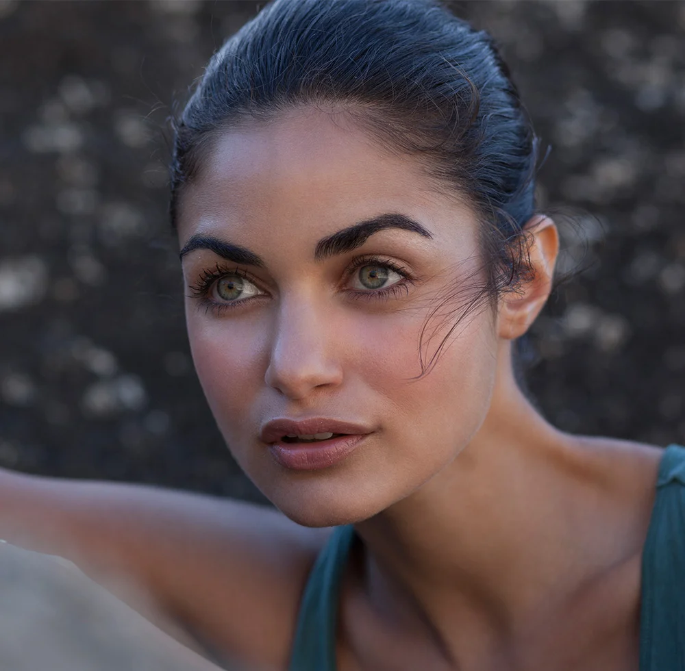 Close-up of a woman with dark hair in a bun, light-medium skin, and green eyes. She wears a teal tank top and looks off into the distance with a neutral expression. Soft light highlights her face against a blurred, stone-textured background. - Skin Pinch Lower Blepharoplasty in Draper, UT
