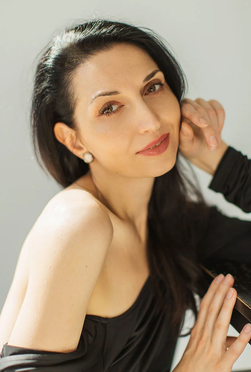 Close-up of a woman with long dark hair, light skin, and a black off-the-shoulder top. She rests one hand on her cheek and the other on a dark surface, looking toward the camera with a pleasant expression. Subtle makeup, pearl earrings, and soft natural light stand out against a neutral gray background. - Skin Pinch Lower Blepharoplasty in Draper, UT