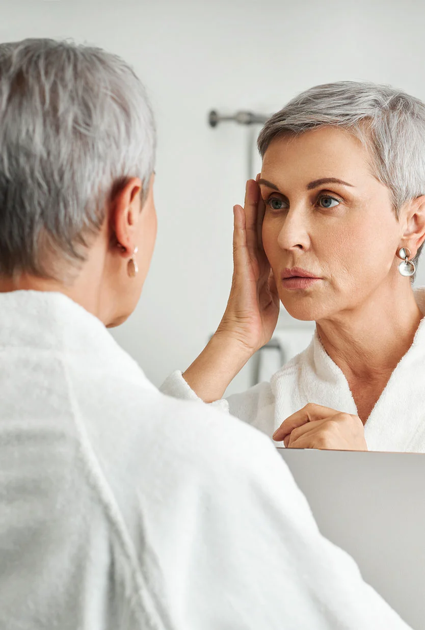A woman with short, gray hair is looking at her reflection in a mirror. She is wearing a white bathrobe. She is touching her face, and her expression appears somewhat concerned or pensive. The lighting in the bathroom is bright and neutral. The background is a light gray/white bathroom wall. The reflection in the mirror shows a similar-aged woman with a similar expression. - Upper Eyelid Surgery in Draper, UT